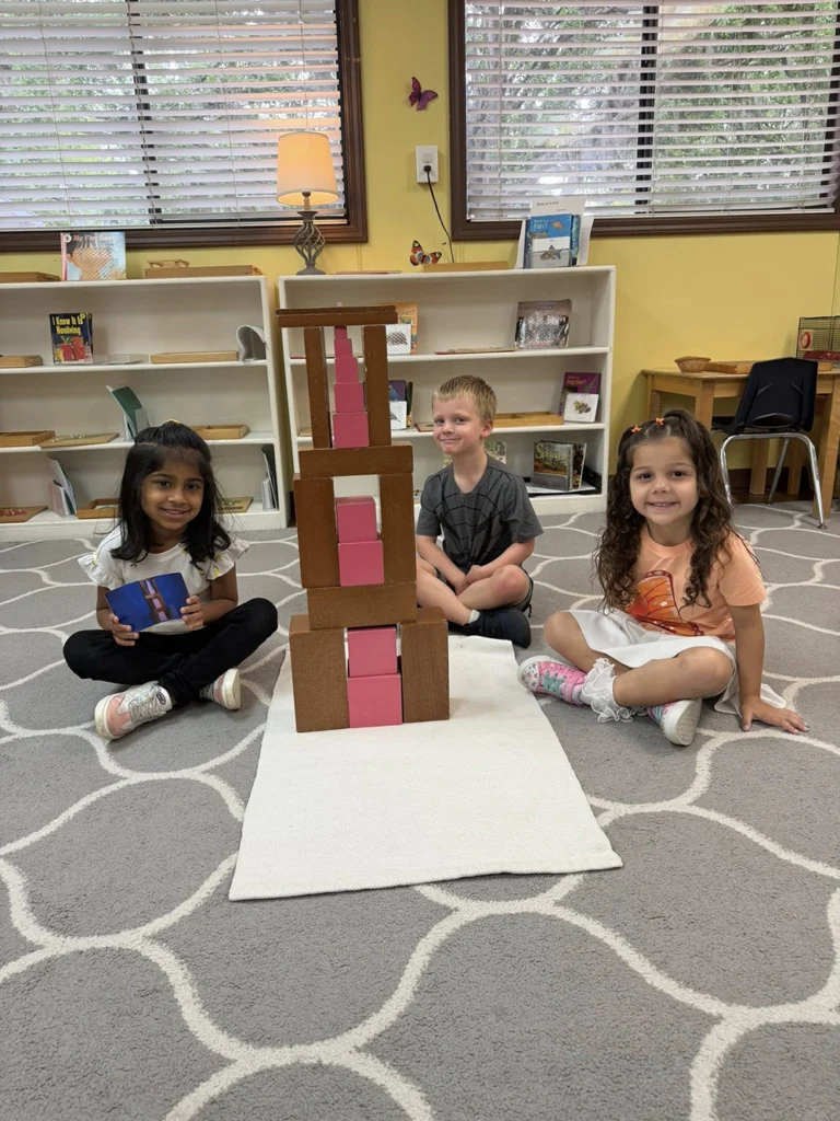 Three Montessori students sitting beside a tower built with brown stair and pink tower materials