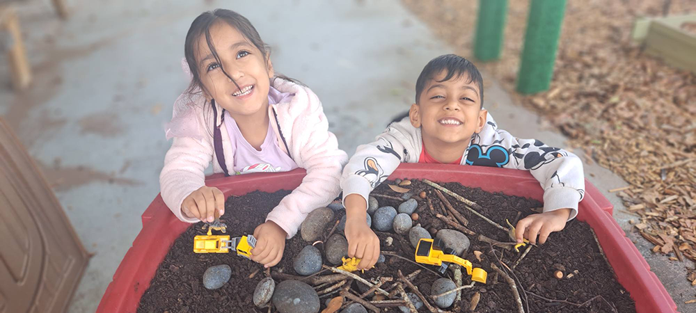 Children smiling while playing with toy trucks in an outdoor sensory table