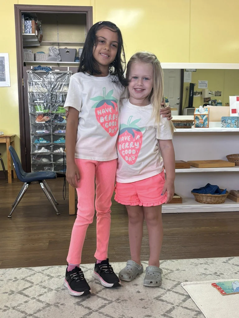 Two children smiling together in a Montessori classroom wearing matching strawberry-themed outfits.